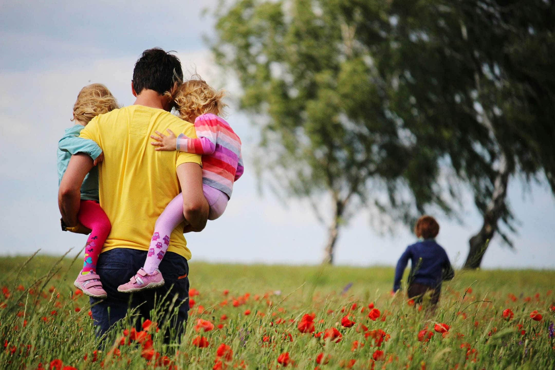 Father walking through a field holding two children whilst the third is running ahead